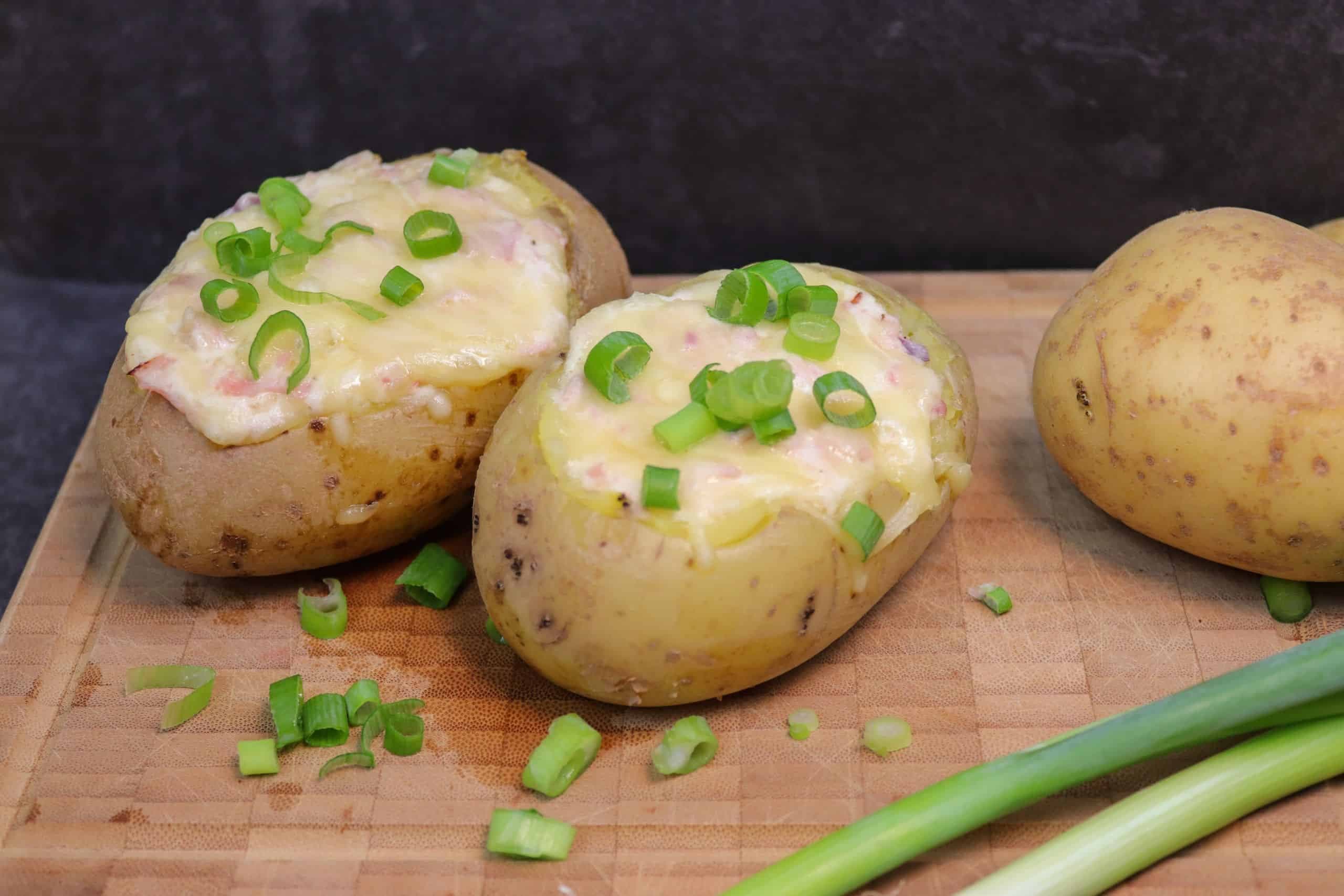 Zwei leckere gefüllte Ofenkartoffeln bestreut mir Lauchzwiebeln auf einem Brett, daneben eine ungekochte Kartoffel und zwei Lauchzwiebeln.