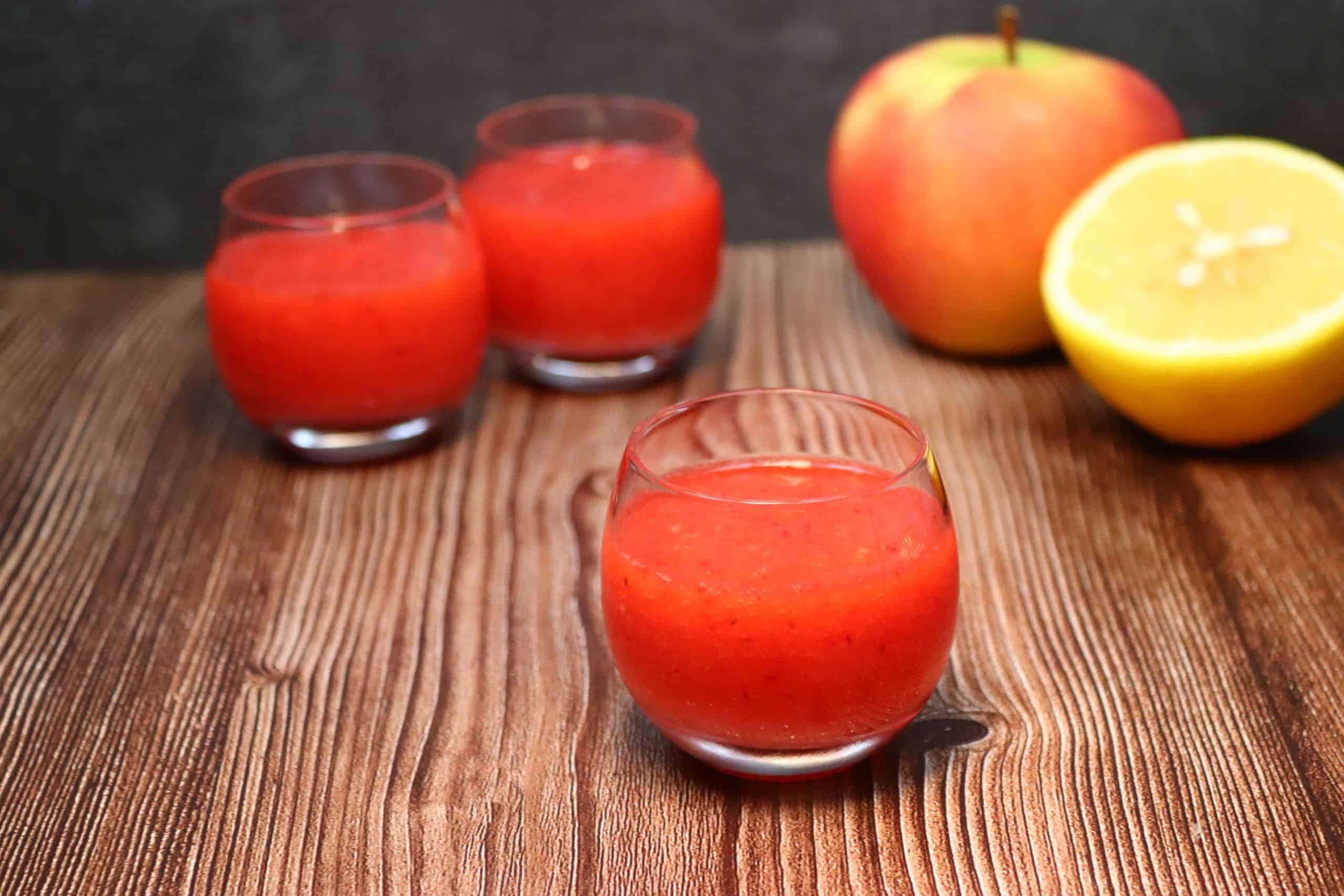 Drei leckere Erdbeer-Vitaminshots mit einem Apfel und einer halben Zitrone auf einem Tisch.