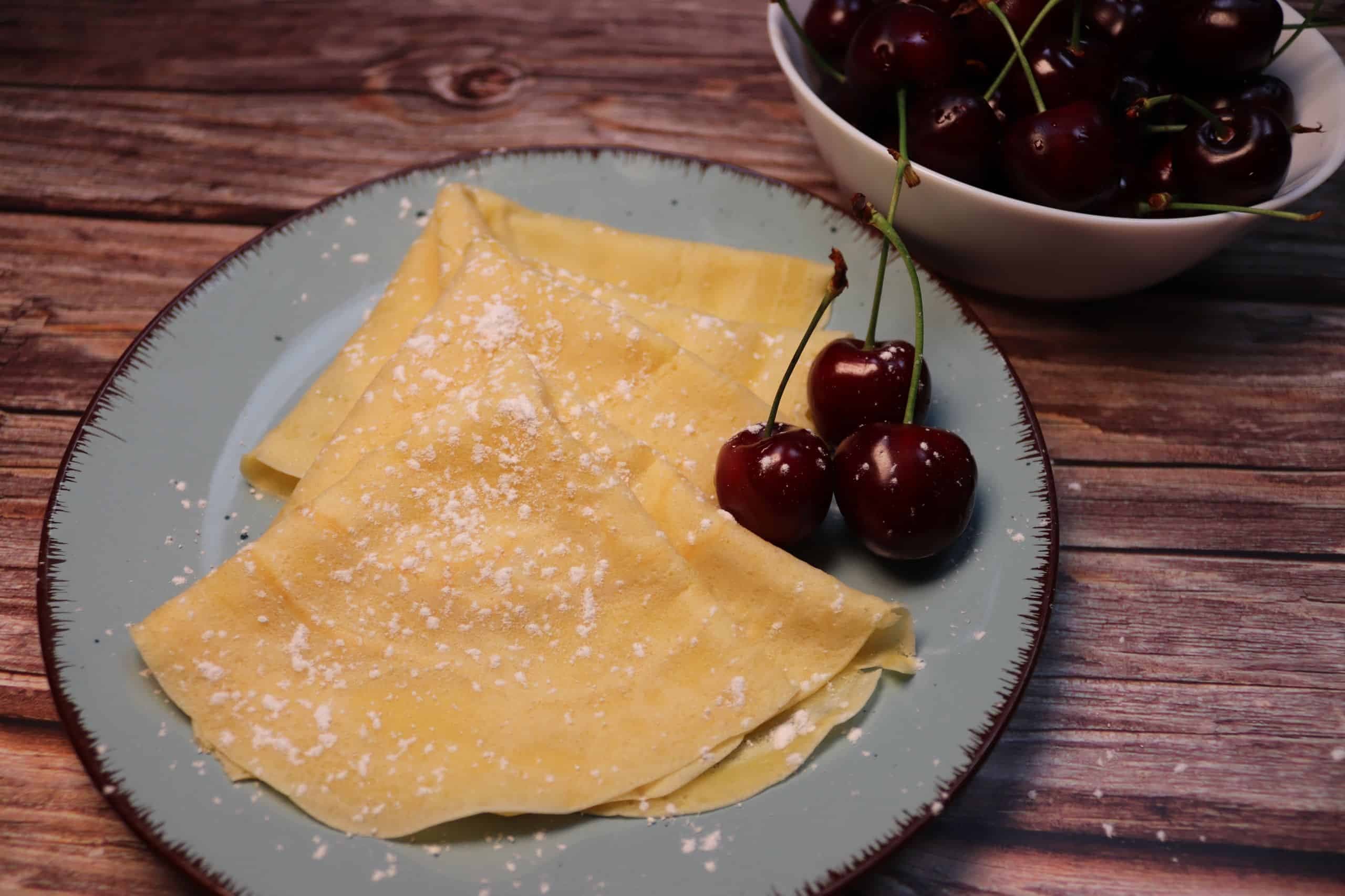 Leckere Crêpes mit Kirschen auf einem Teller, daneben eine Schüssel mit Kirschen